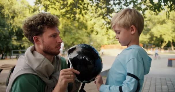 一位满头卷发 留着绿色T恤的父亲帮儿子穿上蓝色毛衣 戴上黑色头盔 然后在公园的一个滑板上滑行 金发小男孩准备骑马 — 图库视频影像
