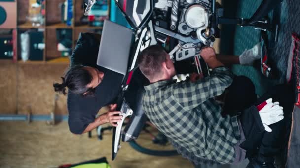 垂直录像 2名机械师修理一辆自行车 一位穿着灰色T恤的黑发男子看着一台灰色笔记本电脑的屏幕 一位穿着格子衬衫的男子在修车厂修理摩托车 — 图库视频影像
