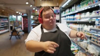 Aşırı kilolu beyaz tişörtü ve kırmızı kulaklıklı siyah önlüklü mutlu erkek süpermarket çalışanı süpermarkette süt koridorlarında dans etmekten ve yürümekten mutlu..