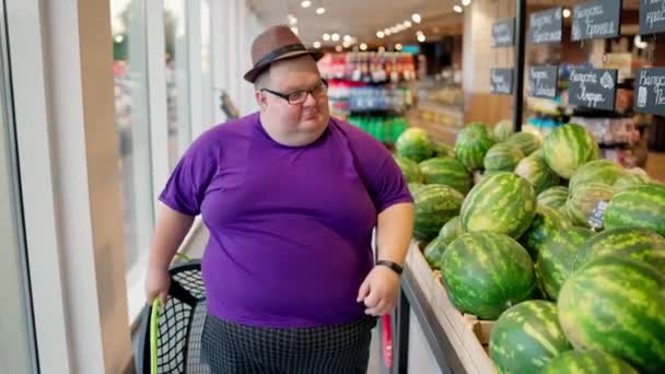 紫色のTシャツと茶色の帽子をかぶった太りすぎの男性が大きなスーパーマーケットでスイカの列に沿って歩き それらを見る — ストック動画
