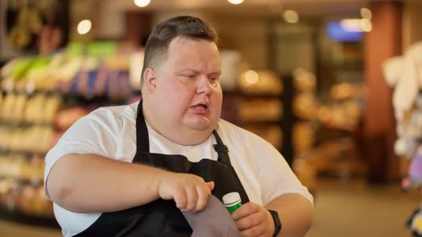 一名身穿白色T恤和黑色围裙的疲惫的男性超市店员在工作辛苦的一天 喝着一瓶绿瓶中的水 擦着额头上的汗珠 — 图库视频影像