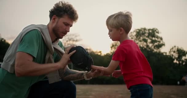 爸爸卷曲的头发 留着绿色的T恤 帮他的小儿子布伦迪穿上红色T恤 戴上棒球手套 在公园里对他拳打脚踢 — 图库视频影像