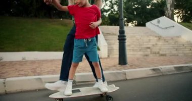 Mavi kotlu bir babanın kırmızı tişörtlü oğluna parkta kaykay sürerken yardım ettiği yakın plan bir fotoğraf. Küçük çocuk parkta beyaz bir kaykaya biniyor..