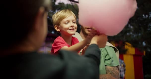 一个穿着红色T恤的金发小男孩 穿着绿色T恤 坐在父亲的怀里 从游乐场收银员手里接过一大块棉花糖 — 图库视频影像