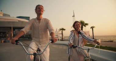 Mutlu bir çiftin önü, bir erkek ve bir kız, yazın güneş doğarken deniz kenarında yetişen palmiye ağaçlarının olduğu plaj boyunca bisiklet sürerler. Güzel modern plaj, erkek ve kız bisiklete biniyor..