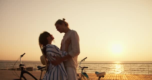 ハッピーカップルと少女は 夏の日の出に海の近くのビーチで抱き合っています 明るい服を着た男と白と青のシャツの女の子が自転車でビーチに到着した — ストック動画