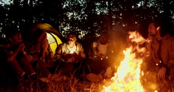 在篝火边 一个小组坐在炉火边 在地板和秋夜的森林里讲述着恐怖的故事 戴帽子的男人会亮起手电筒 — 图库视频影像