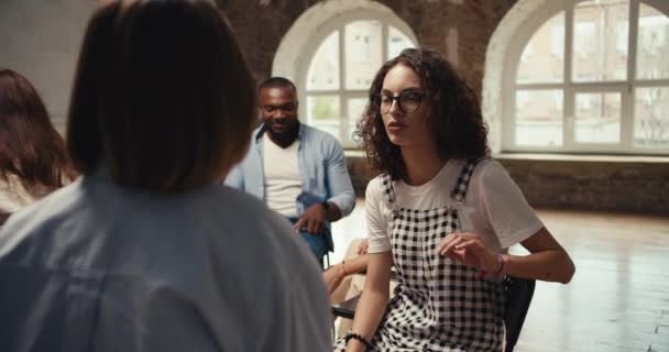 メガネのカーリーヘアとチェッカーのジャンプスーツを持つブルネットの少女は 茶色のホールでのグループ療法中にペアで通信します — ストック動画
