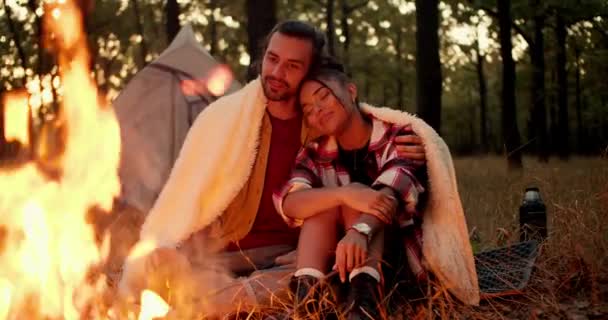 一个黑头发的男人和一个黑皮肤的女孩在篝火边的白色毛毯下 靠着绿色森林里的帐篷 一边坐着一边拥抱 愉快舒适的夜晚在休息时 — 图库视频影像