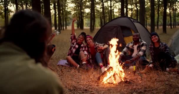 一名身穿绿色夹克的黑发男子在火堆边穿着格子衬衫拍摄了一群徒步旅行者的照片 一个人在绿色和黄色的秋天的森林里弹奏吉他 人们坐在帐篷旁边的画框里 — 图库视频影像