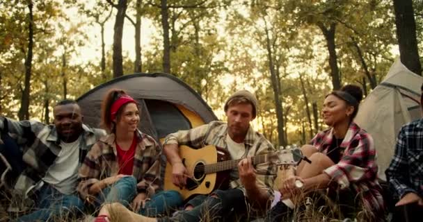 在一个阳光明媚的夏天的森林里 一个金发男子和其他远足的人在一起 一边和他一起弹奏吉他 一边在帐篷的背景下听着他的悦耳的吉他音乐 — 图库视频影像