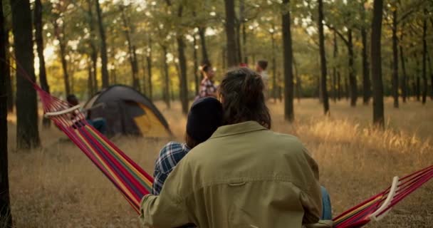 一对快乐的夫妇 一个黑发男人和一个戴着黑色帽子的女孩 坐在一家公司旁边的红色吊床上 在阳光灿烂的夏天的森林里露营 — 图库视频影像