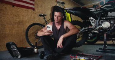 Gri tişörtlü kısa saçlı erkek tamirci, bir mola sırasında atölyede motosiklet tamir ederken beyaz telefonuyla çalışıyor. Bir adam atölyesinde aletlerin yanında oturuyor..