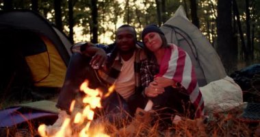 Siyah tenli bir adam ve siyah şapkalı bir kız kamp sırasında ateşin yanında oturur ve güzel bir ormanın arka planında Amerikan bayrağının altında çadır kurar..