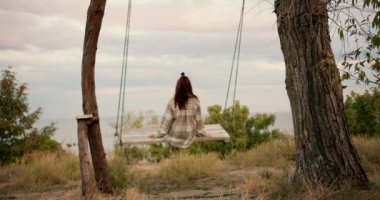 Arkasından ateş eden: Kareli tişörtlü esmer bir kız şehrin dışındaki ahşap salıncakta sallanıyor. Denizin kıyısında dinlenmek.