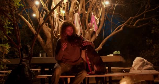 一个卷发男孩在篝火边的长椅上 靠着森林背景喂一只黑狗 在乡间别墅休息 — 图库视频影像