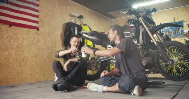 两名快乐的男性技师在车库里的车间里喝着小瓶子里的饮料 在车间工作时休息一下 汽车机械师二重唱 — 图库视频影像