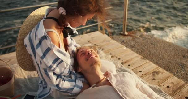 一个穿着浅色衣服的快乐的金发男人躺在他棕色头发的女朋友的膝上 在海边的码头野餐 一个男人和一个女孩在海边放松一下 — 图库视频影像