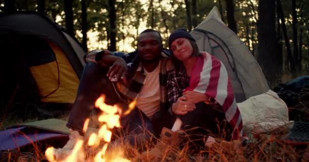 一个黑皮肤的男人和一个戴着黑色帽子的女人坐在篝火边 在美丽的森林和帐篷的背景下 在美国国旗下拥抱 — 图库视频影像