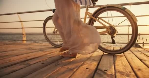 一个身穿白色长裙的女孩的特写镜头 她在风中扭动着身子 在海滩边走着自行车 海滩上的木板上覆盖着夏天的日出 — 图库视频影像