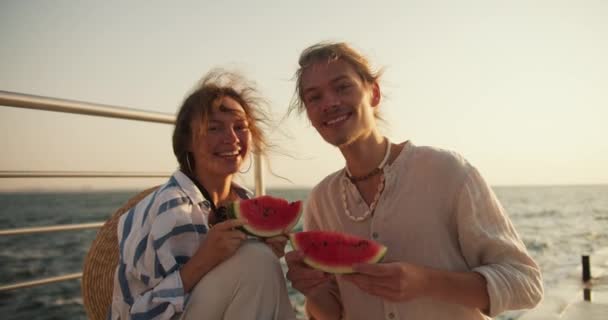 一对快乐的夫妇的画像 一个穿着浅色衣服的男人和一个穿着蓝色和白色衬衫的女孩 手里拿着几片西瓜 看着摄像机 男人和女孩在码头野餐 — 图库视频影像