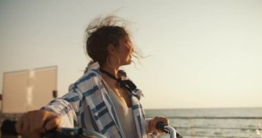 Mutlu kahverengi saçlı bir kız deniz boyunca sahil boyunca yürür ve bisikletini yanında taşır. Deniz kenarında bisiklet süren bir kız..