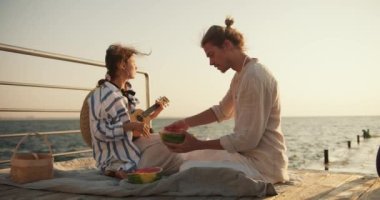Bir adam ve bir kız iskelede oturuyorlar, açık renk elbiseli bir adam karpuz kesiyor ve bir kız ukulele çalıyor ve denize bakıyorlar. Mavi tişörtlü bir kız bir müzikalde çalıp şarkı söylüyor.