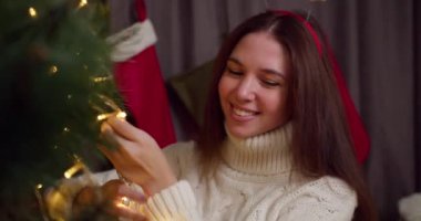 Beyaz kazaklı mutlu esmer bir kızın yakın çekimi, yeni yıl ağacını kışın sıcacık bir evde parıldayan sarı bir çelengin yardımıyla süslüyor ve yeni yıl tatiline hazırlanıyor.