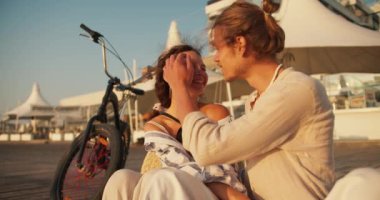 Bir randevuda mutlu bir çift, açık renk elbiseli sarışın bir adam kumsalda bisikletin yanında oturan açık renk elbiseli, siyah saçlı bir kızın saçını düzeltiyor..