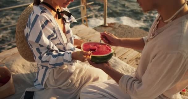 一个男的和一个女的在海边的一个码头野餐时 带着勺子吃西瓜 一对快乐的夫妇 一个金发碧眼的男人 一个棕色头发的女孩 穿着白色的蓝色衬衫 坐在海边的一个码头上 — 图库视频影像