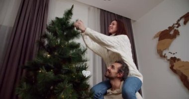 Mutlu çift erkek ve kız yılbaşı ağacını süslüyor, erkek, kız arkadaşının omuzlarına oturmasına yardım ediyor ve kışın rahat bir evde yeşil Noel ağacının tepesine yeni yıl oyuncağını asıyor..