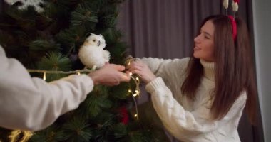 Beyaz kazaklı esmer bir kızın yakın plan fotoğrafı, erkek arkadaşıyla birlikte, Yeni Yıl ağacını süsleyin ve parlak sarı bir çelenk yardımıyla kışın Noel için hazırlanın.