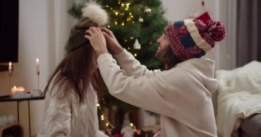 Yeni yıl şapkalı mutlu bir adam beyaz kazaklı esmer kız arkadaşının kafasına yeşil-kahverengi bir şapka takıyor. Birbirlerine bakıyorlar ve yeni yıl ağacının yanında gülümsüyorlar.