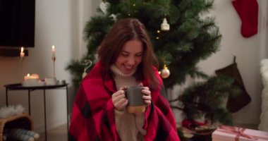 Kırmızı kareli ekose gömlekli mutlu esmer kız gri bardaktan sıcak çay içiyor, gülümsüyor ve kışın sıcak bir odada Noel ağacının yanına bakıyor..