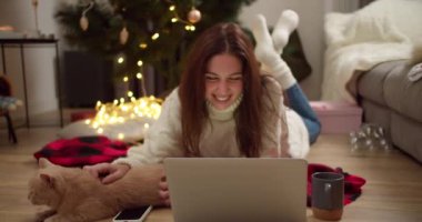Beyaz kazaklı mutlu esmer kız kırmızı bir ekoseyle yere uzanıp dizüstü bilgisayarda film izliyor ve Noel tarzı dekore edilmiş rahat bir odada krem rengi kedisini okşuyor..