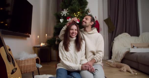 一对快乐的夫妇的画像 一个穿着白色毛衣的黑发男人和一个穿着白色毛衣的黑发女孩坐在圣诞树旁的地板上 他们的奶油色猫躺在舒适的环境中 — 图库视频影像