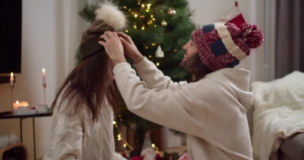 新しい年の暖かい帽子の幸せな男は 白いセーターに彼のブルネットガールフレンドの頭の上に緑色の茶色の帽子を置きます 彼らはお互いを見て 大気中の新年の木の近くで微笑みます — ストック動画