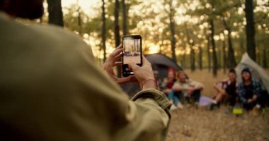 Açık yeşil tişörtlü bir adam, güneşli yeşil bir yaz ormanında telefonundaki kamerayla yürüyüş yaptığı şirketinin fotoğraflarını çekiyor. Kamp durağında hatıra olarak fotoğraf çekiyorum..