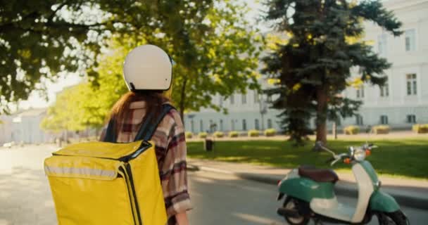 一个身穿格子衬衫 头戴白色头盔 头戴黄色大包的女信使坐着摩托车走到她身边的后视镜 她骑上摩托车 在阳光灿烂的情况下开始在公园附近的街上点菜 — 图库视频影像