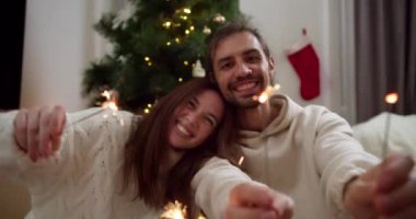 Mutlu bir çiftin portresi, beyaz kazaklı bir erkek ve kız ellerinde maytap, yaklaşan Noel tatili için seviniyorlar ve dekore edilmiş bir evin sıcak atmosferinde eğleniyorlar.