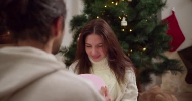 Omzunun üzerinden, bir erkek kız arkadaşına pembe bir hediye verir ve kutuyu kaldırır, kız hediyeye bakar ve sonra adama bakar ve yeni yıl ağacının yanında rahat bir atmosferde geniş bir gülümseme sergiler.