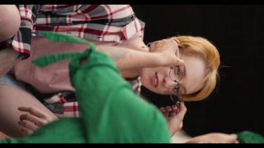 Dikey video: yeşil saçlı bir kızın kız arkadaşına gözlüğünü çıkartırken, kısa saçlı bir sarışının kareli pembe gömlek giyip randevusunda onunla konuşurken yakın plan fotoğrafı..