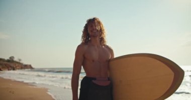 Dalgalı mutlu bir sörfçünün portresi. Çıplak bir gövdesi var. Sörfü elinde tutuyor ve kumsalda duruyor..
