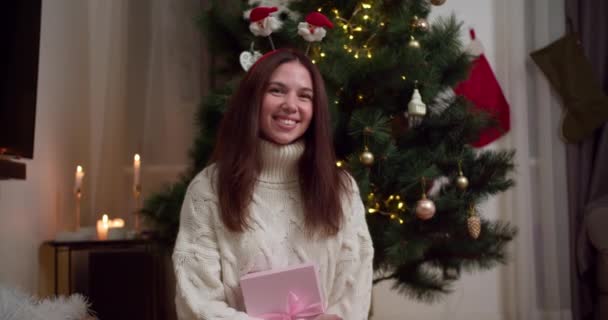 一个穿着白色毛衣的快乐的黑发女孩的画像 她在一个舒适的冬天的家 在一棵装饰过的圣诞树旁拿着一个粉色礼品盒 — 图库视频影像