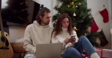 Ciddi ve düşünceli esmer bir adam ve beyaz kazaklı bir kız yerde oturup yeni yıl ağacının yanında bir dizüstü bilgisayar ve telefon üzerinde çalışırlar..