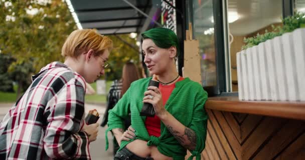 在约会期间 两个穿着格子呢衬衫 留着短发的女孩在公园的一家街头咖啡馆边吃热狗 并进行交流 — 图库视频影像