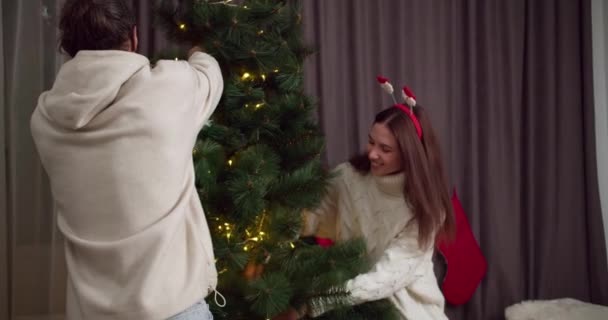 A brunette guy in a white sweatshirt together with his brunette girlfriend in a white sweater decorate the Christmas tree and prepare for the Christmas holiday in their cozy home in winter. Happy