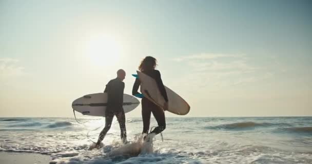 在一个阳光明媚的夏日早晨 两个穿着湿衣的冲浪男手拿着冲浪板走进了水里 两个男人夏天在海滩上冲浪 — 图库视频影像