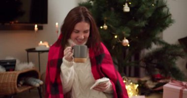 Kırmızı ve beyaz kazaklı mutlu esmer kız gri bardaktan sıcak çay içer ve Noel ağacının yanındaki süslü bir odada beyaz telefonuyla çalışır..