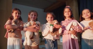 Beş anaokulu öğrencisinin portresi. Yumuşak oyuncakları duruyor, kameraya bakıyor ve çocukları okula hazırlamak için bir kulüp odasında gülümsüyor..